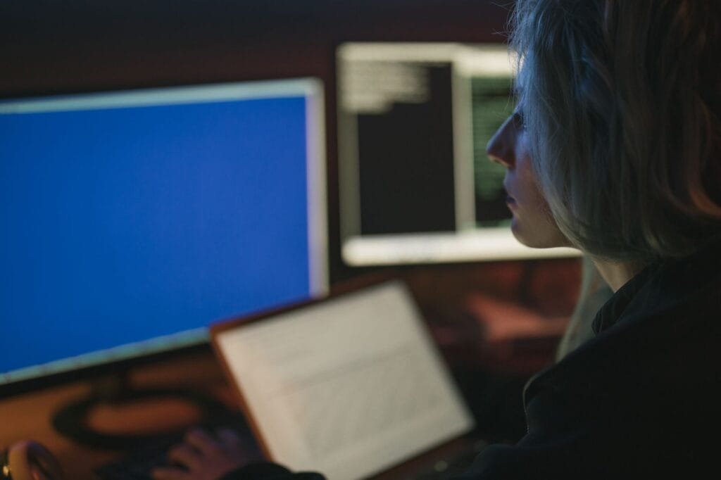 During a cybersecurity solutions review, an analyst studies code on multiple monitors while checking system activity in a dim workspace.