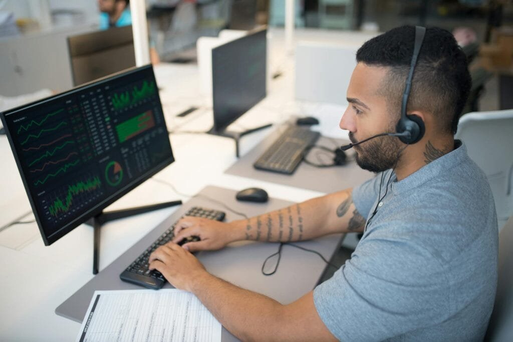 A cybersecurity analyst monitors threat dashboards on a desktop screen while wearing a headset and typing at an office workstation.